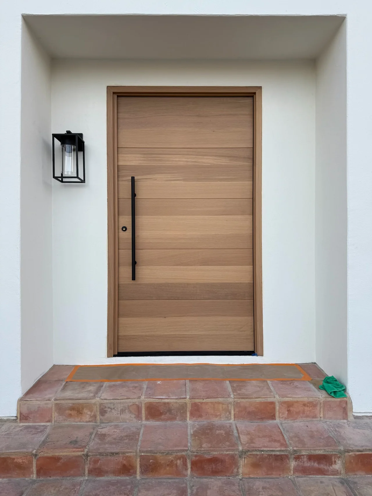 Natural wood entry door with a long black handle beside a black lantern in a white stucco entrance